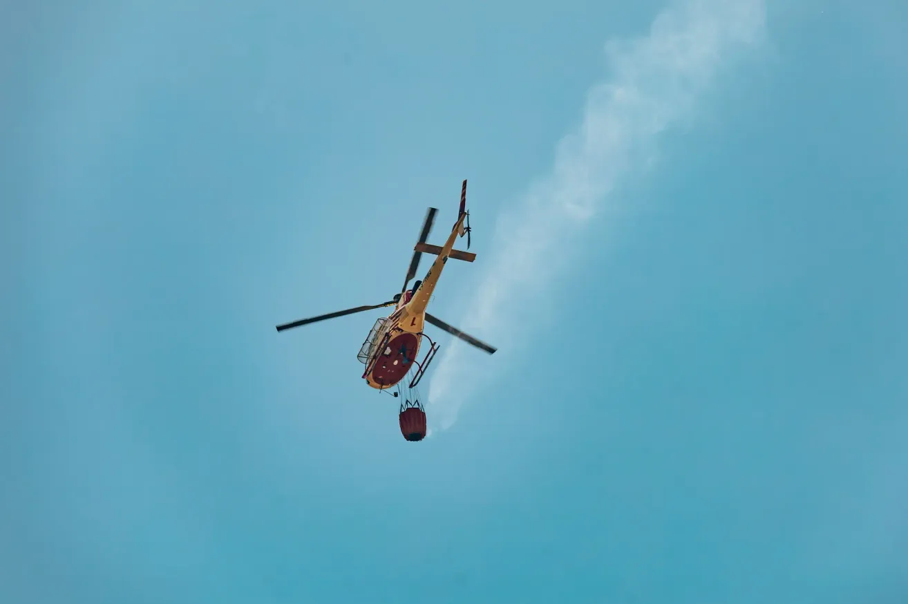 A yellow helicopter with a red bucket is flying in the sky, likely used for firefighting.