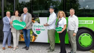 Five people stand in front of a green bus, holding green heart-shaped signs. They are smiling and appear to be posing for a photo. The bus has a white and green design with the text Einfach kostbar.