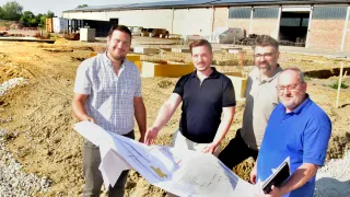 Four men stand on a construction site, holding a large blueprint. They are smiling and appear to be discussing the plans. Behind them, a building with solar panels is visible.