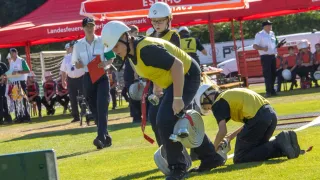 A group of firefighters are practicing on a field. One is kneeling, holding a hose, while another stands holding a fire extinguisher. A woman in the background holds a clipboard.