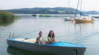 Two women are sitting in a blue and white boat on a lake with mountains in the background.