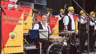 A man in a white shirt and black vest plays drums on a stage with a band in the background.