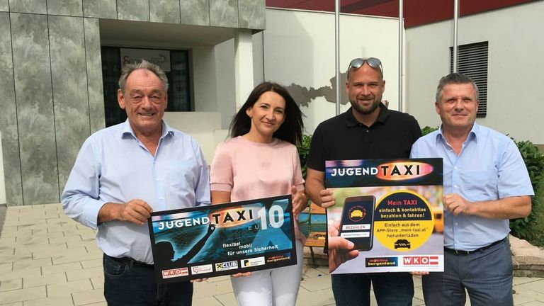 Three people stand in front of a building, smiling and holding signs. The man on the left holds a sign with 'JUGENDTAXI 10 EURO'. The woman in the middle holds a sign with 'flexible mobil für unsere Sicherheit'. The man on the right holds a sign with 'JUGENDTAXI' and a smartphone.