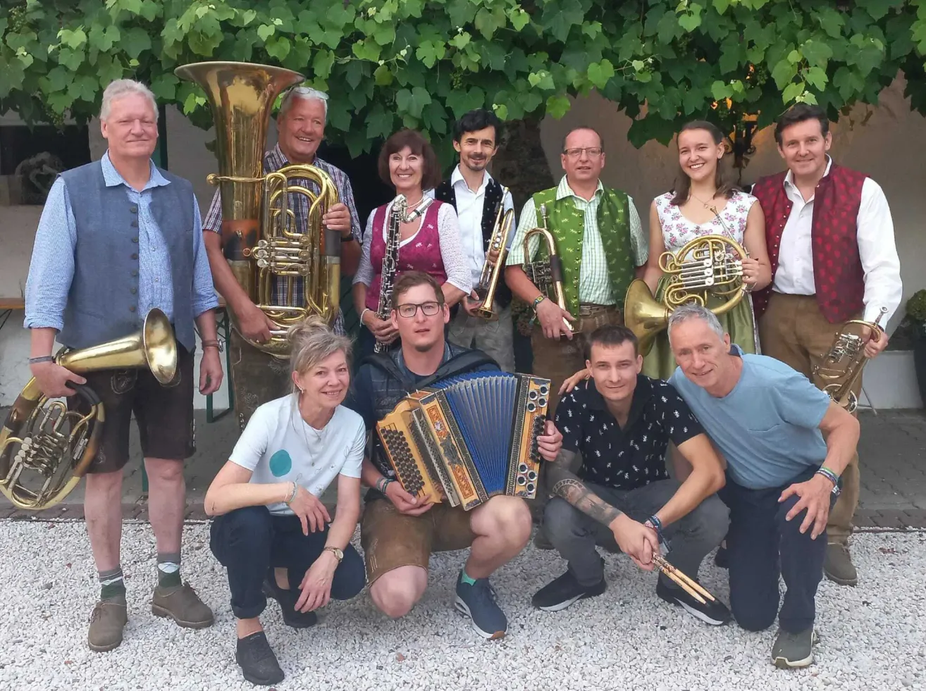 A group of musicians, including brass and woodwind instruments, are posing for a photo in front of a green leafy backdrop.
