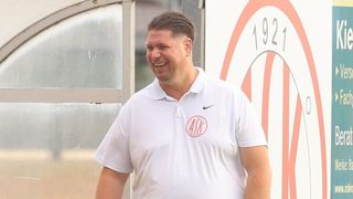 A man wearing a white Nike polo shirt with a red ASK logo on the left chest area, smiling and standing in front of a wall with a red circle and the number 192 on it.