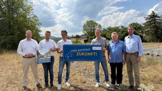 Five men stand in a field, holding a blue sign that reads 'Hier entsteht Zukunft!' with a megaphone icon. They are dressed casually and appear to be posing for a photo.