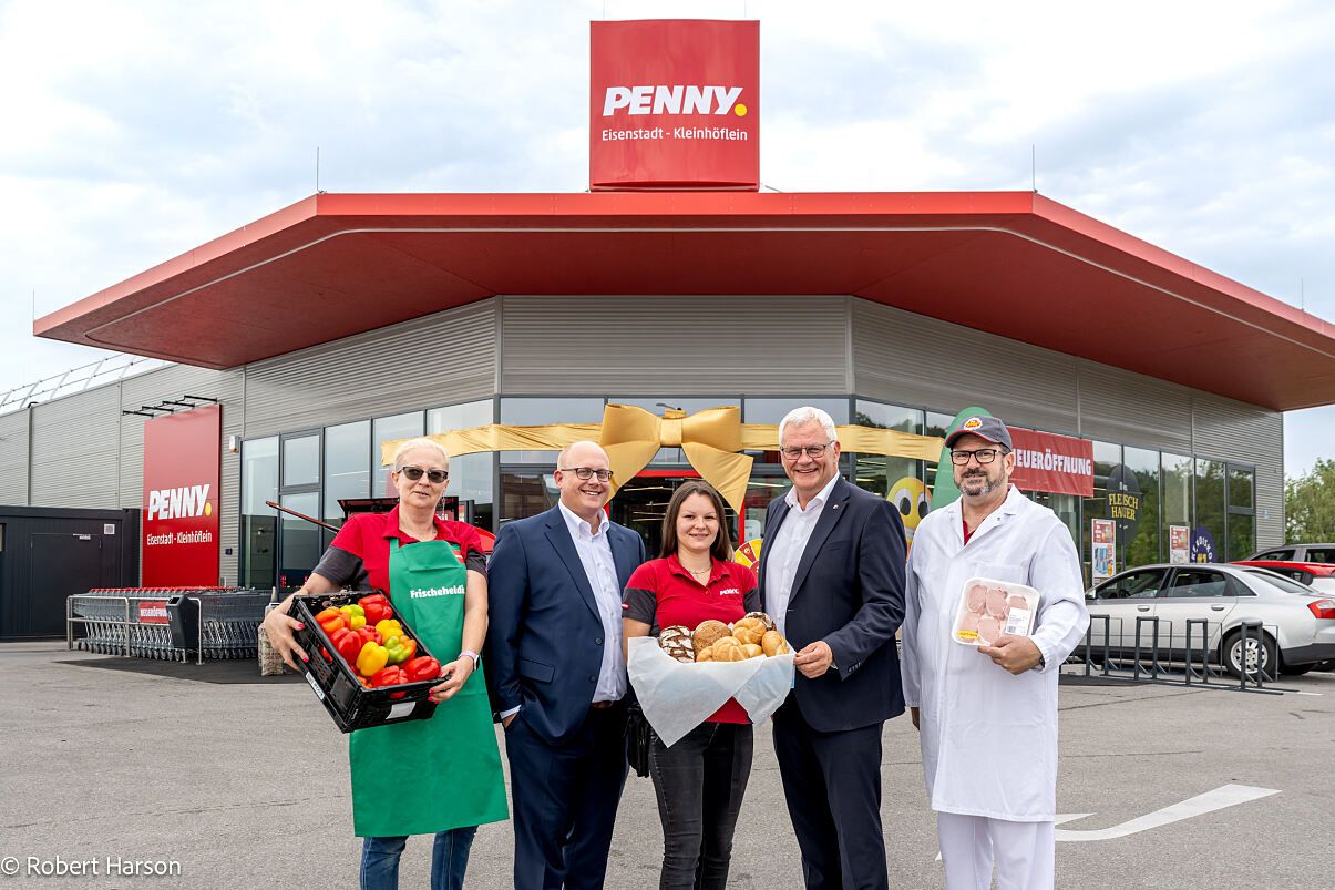 Eine Gruppe von Menschen steht vor einem PENNY-Laden, jeder hält einen Korb mit Essen. Die Frau in der Mitte hält einen Korb mit Brot und Gebäck. Der Mann auf der rechten Seite hält eine Packung Fleisch. Die Frau auf der linken Seite hält einen Korb mit Gemüse. Der Laden hat ein rotes Dach und ein Schild, das PENNY steht.