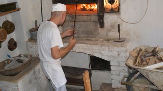 A man in a white apron and hat is using a long stick to tend to a fire in a brick oven.