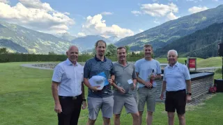 Fünf Männer stehen auf einem Golfplatz mit Bergen im Hintergrund. Sie lächeln und halten Trophäen und Flaschen.