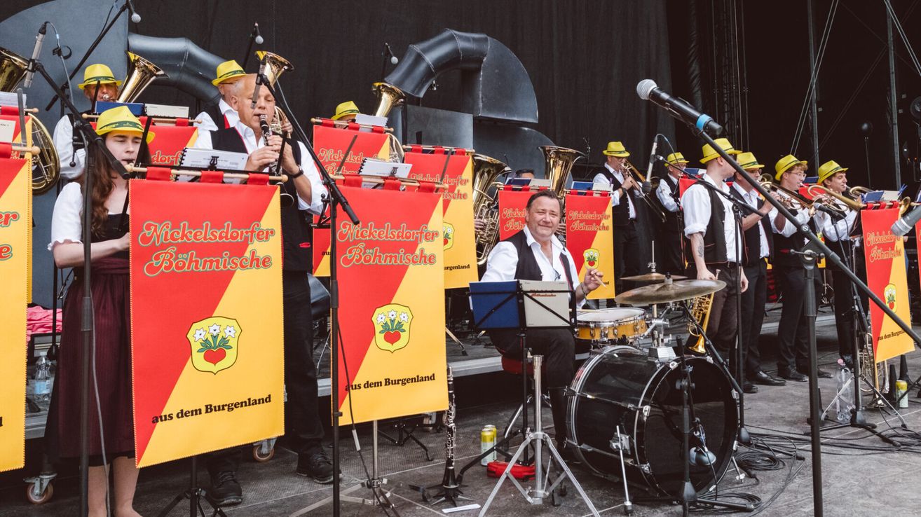 Eine Band von Musikern, einschließlich eines Mannes, der Klarinette spielt, tritt auf der Bühne auf, mit einem Mikrofon, Trommeln und anderen Instrumenten. Sie tragen gelbe Hüte und weiße Hemden.