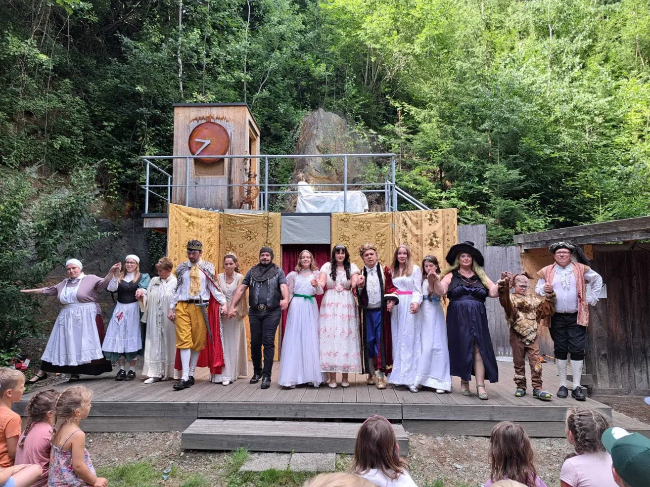 A group of people dressed in medieval costumes stand on a wooden stage in a forest setting, with a backdrop of trees and a small wooden structure with a red circle.