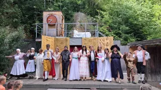A group of people dressed in medieval costumes stand on a wooden stage in a forest setting, with a backdrop of trees and a small wooden structure with a red circle.