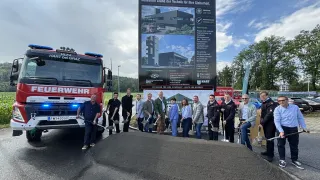 A group of people in formal attire are gathered around a large signboard, holding shovels and posing for a photo. A fire truck is parked on the left side, and a fireman is standing in front of it. The signboard has a picture of a building and some text.