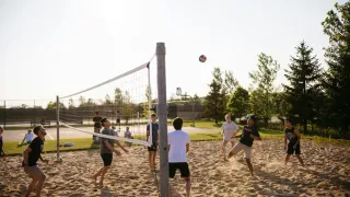 A group of people are playing volleyball on a sandy court. The ball is in the air, and the players are in various positions.