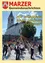 Eine große Gruppe von Menschen versammelt sich um einen Maibaum vor einer Kirche, während eine Menge zuschaut. Das Bild hat einen gelben Rand und enthält Text auf Deutsch.