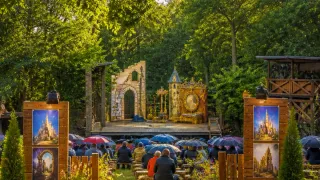 A group of people are seated under umbrellas in an outdoor theater setting, watching a performance on a stage with a backdrop of a castle and a clock tower.