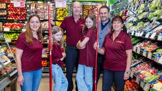 A family of five, including two young girls, stand together in a grocery store, all wearing matching maroon shirts. Behind them, shelves are stocked with various fruits and vegetables.