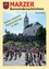 Eine große Menschenmenge versammelt sich um einen Maibaum vor einer Kirche und feiert ein traditionelles Fest. Die Kirche ist mit einem Glockenturm geschmückt und von Bäumen und einer Steinmauer umgeben.