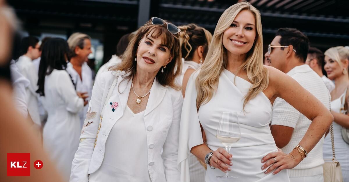 Two women in white outfits, one with a pearl necklace and sunglasses on her head, and the other holding a wine glass, smiling and standing together in a crowd.