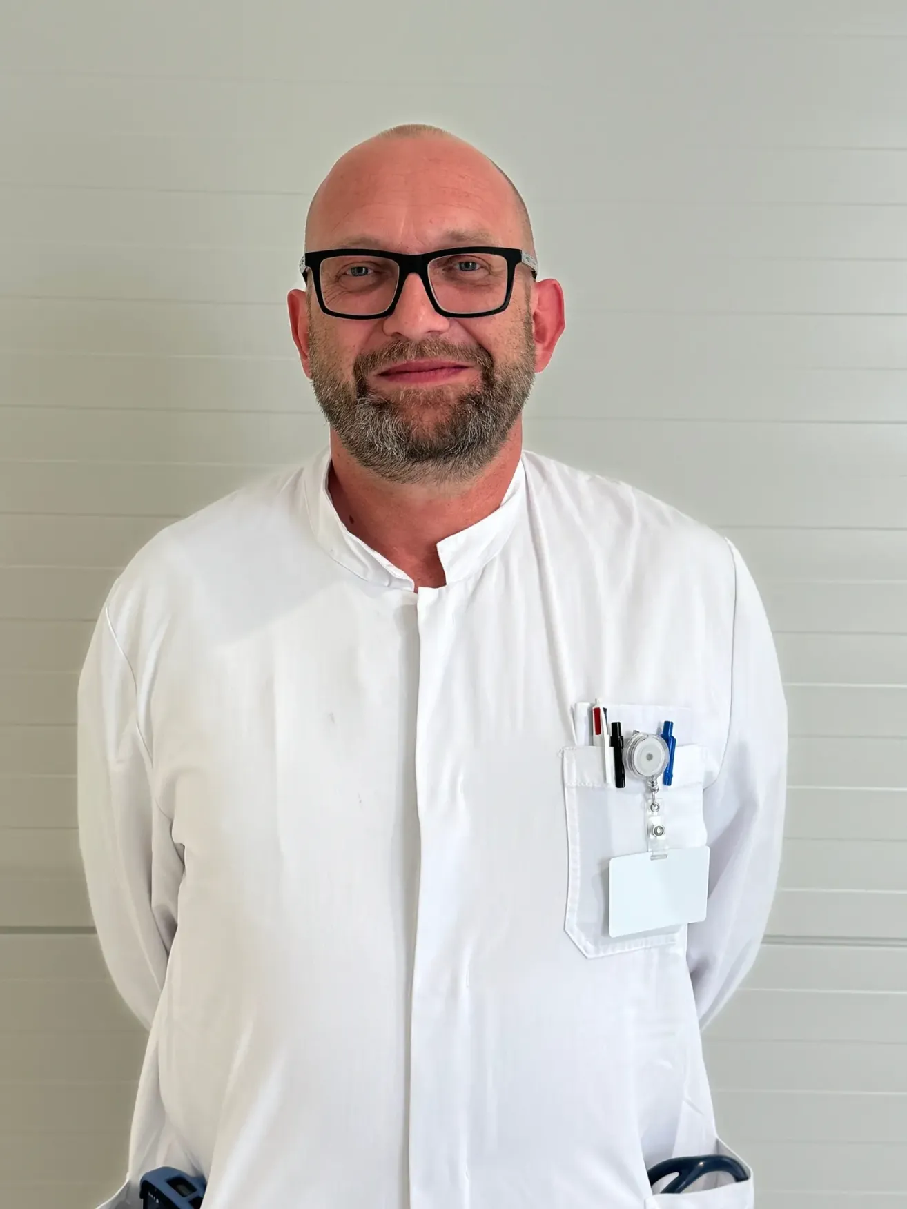 A man in a white lab coat stands against a white wall, smiling and looking forward. He wears glasses and has a badge and pen in his pocket.