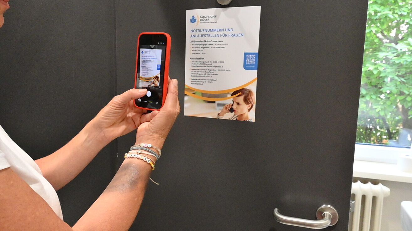 Eine Person hält ein rotes Smartphone und fotografiert ein Poster mit Notfallnummern für Frauen.