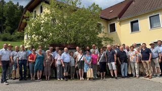 A group of people in casual attire, smiling and posing for a photo in front of a yellow house with a tree in the center.