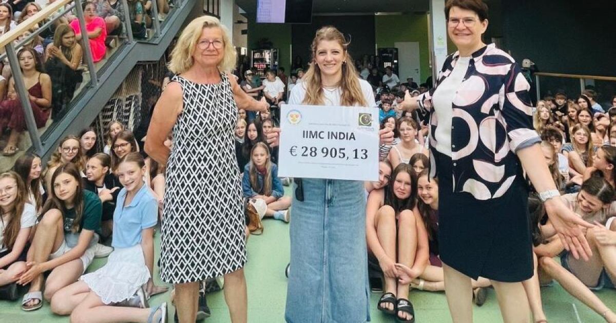 Three women stand in front of a crowd holding a sign with a large amount of money. The woman in the middle holds a certificate with the text 'IMC INDIA' and the amount '€ 28,905.13'.