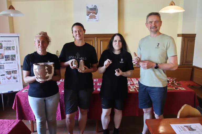 Four people stand in a room, holding various clay objects. Two women hold clay pots, one man holds clay pieces, and another woman holds a clay figurine. A table with items is behind them.