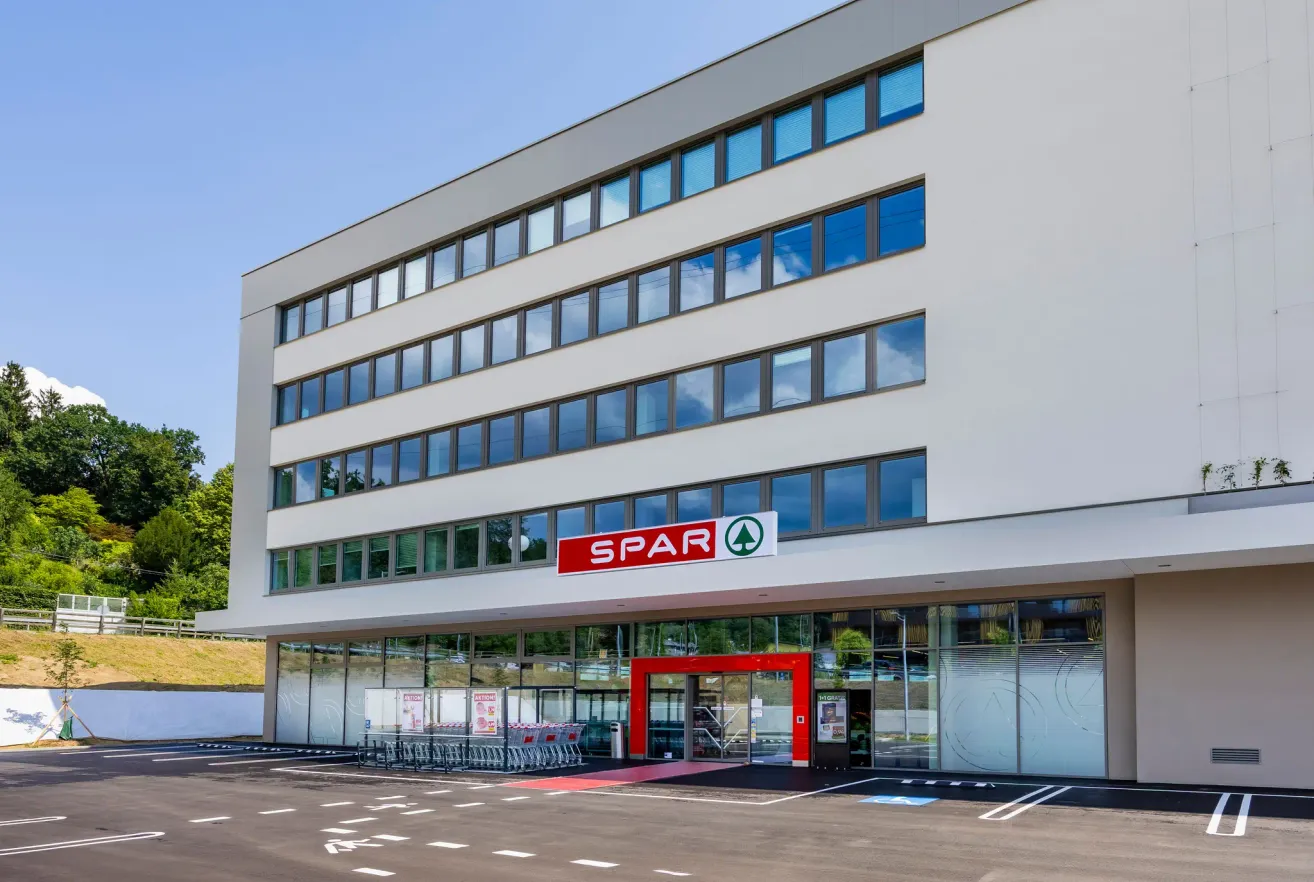 A modern office building with a red entrance and a sign that reads SPAR. The building has many glass windows and a parking lot in front.