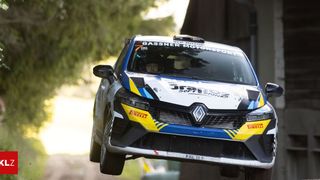 A rally car, sponsored by Gassner Motorsport, is airborne in a jump. The car has a yellow and blue livery with the word 'Soft Drinks' on the front.