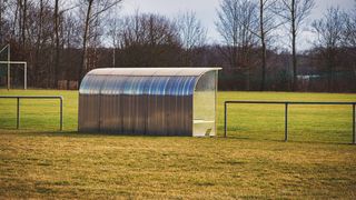 A metal and glass structure stands on a grassy field with a metal fence in the foreground and bare trees in the background.