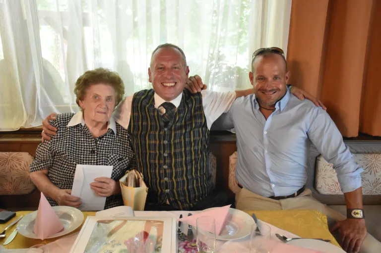 Drei Menschen sitzen an einem Tisch und lächeln für ein Foto. Eine Frau hält ein Papier, ein Mann in einem Sakko und ein Mann in einem blauen Hemd. Eine Tischdecke, Teller und Gläser sind auf dem Tisch.