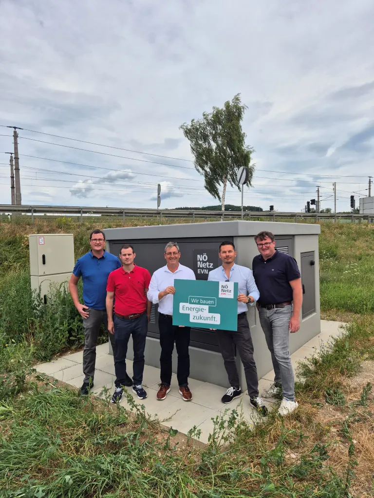 Fünf Männer stehen vor einer großen grauen Box mit den Worten 'Wir bauen Energie-zukunft' auf einem grünen Schild. Sie lächeln und scheinen für ein Foto zu posieren.