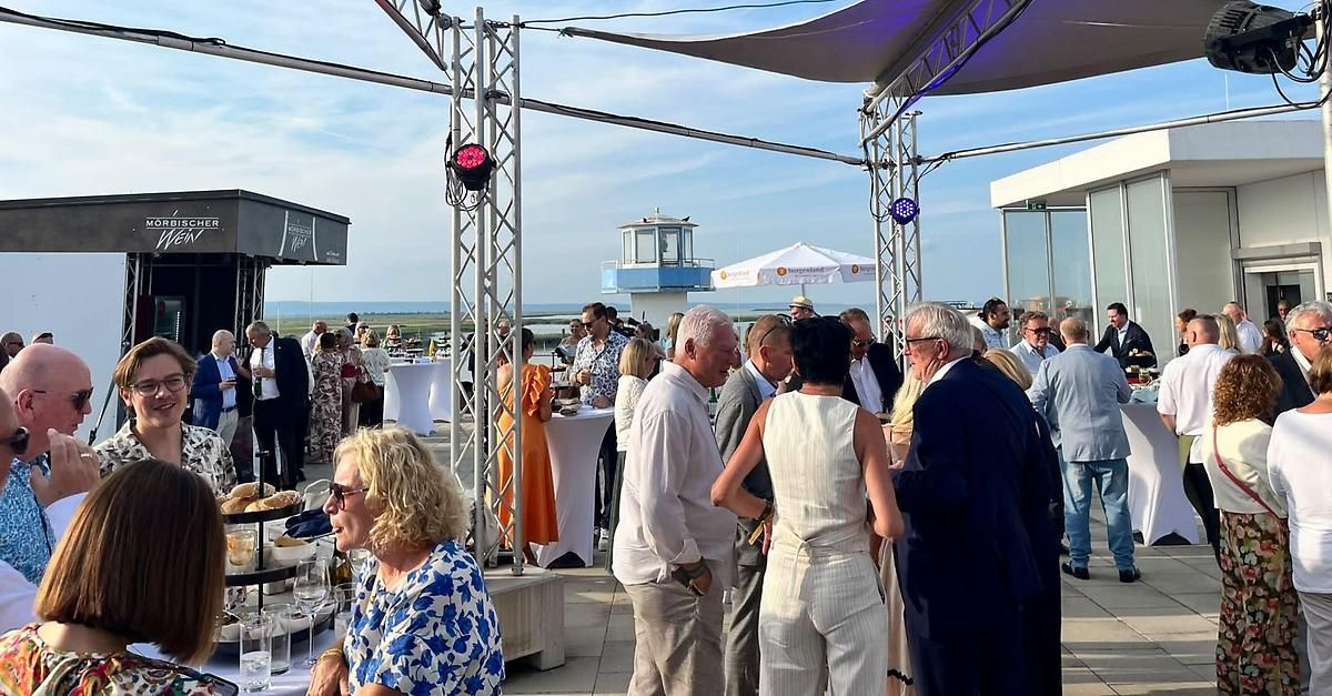 Eine Gruppe von Menschen versammelt sich auf einem Dach, einige stehen und andere sitzen, mit aufgestellten Tischen und Stühlen. Die Umgebung scheint ein Outdoor-Event mit Blick auf das Meer zu sein.