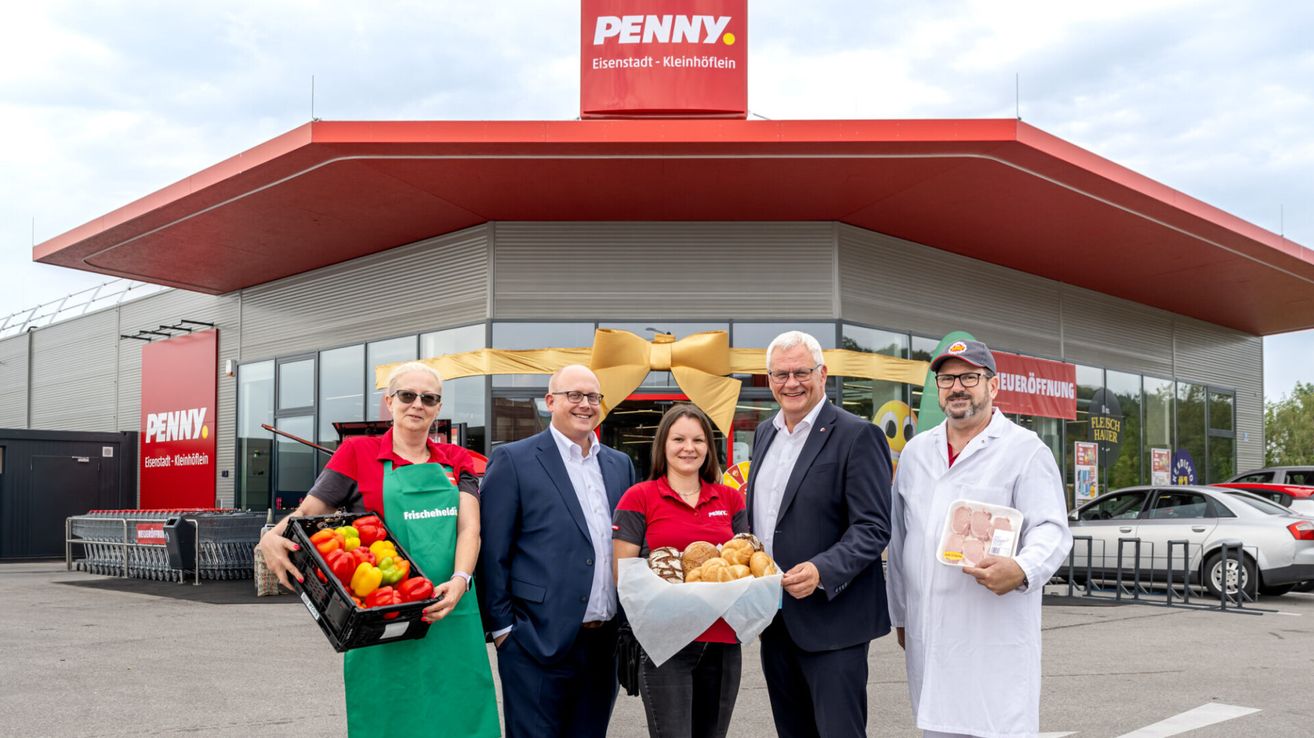 Fünf Personen stehen vor einem PENNY-Geschäft, zwei halten Obst und Gemüse und zwei halten Lebensmittel. Die Frau in der Mitte hält einen Korb mit Brot.