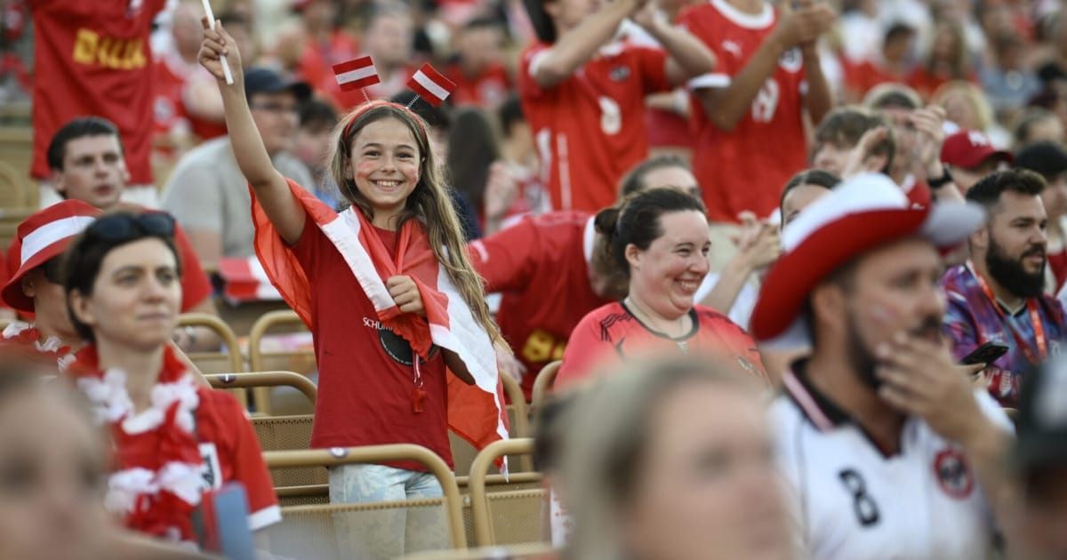 Ein junges Mädchen mit roten Flaggen auf dem Kopf und einem roten Trikot jubelt in einem Stadion mit anderen Fans.