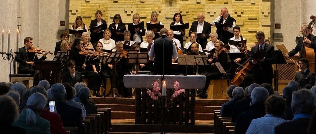 Ein Chor und ein Orchester spielen in einer Kirche, mit Musikern, die Violinen, Violoncelli und andere Instrumente spielen. Der Dirigent steht vor einer Kerze auf einem Ständer, und das Publikum sitzt auf Kirchenbänken.