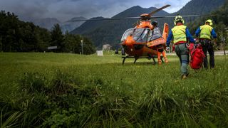 Ein Hubschrauber mit der Aufschrift 'Luftrettung' an der Seite steht auf einem Grasfeld, und eine Person in einer gelben Weste geht darauf zu. Im Hintergrund befinden sich Berge und ein bewölkter Himmel.