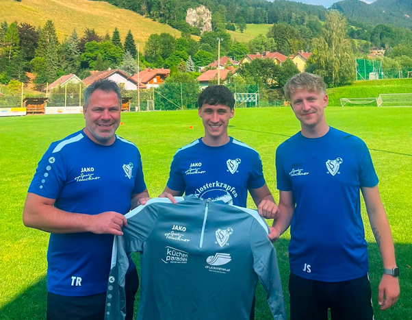 Three men in blue jerseys stand on a grassy field, holding a blue shirt with white text and logos. The man on the left wears a watch, and the man on the right has a tattoo on his arm. Behind them, houses and a mountain range are visible.