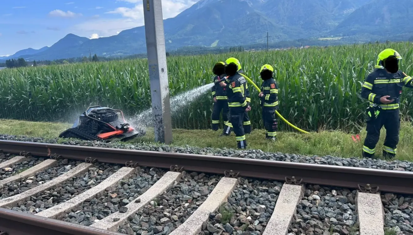 Firefighters in protective gear are using a water sprayer to extinguish a fire near a train track in a cornfield.
