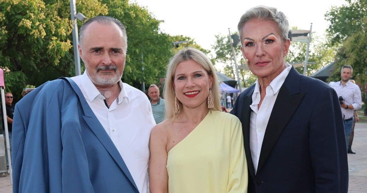 Drei Personen, zwei Frauen und ein Mann, stehen eng beieinander und lächeln für ein Foto. Die Frau links trägt ein gelbes Kleid, während der Mann links einen blauen Anzug trägt. Die Frau rechts trägt einen schwarzen Anzug.