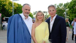 Drei Menschen stehen eng beieinander und lächeln für ein Foto. Der Mann auf der linken Seite trägt eine blaue Jacke, die Frau in der Mitte ein gelbes Kleid und die Frau auf der rechten Seite ein blaues Kostüm. Sie befinden sich in einem Außenbereich mit Bäumen und einem Gebäude im Hintergrund.