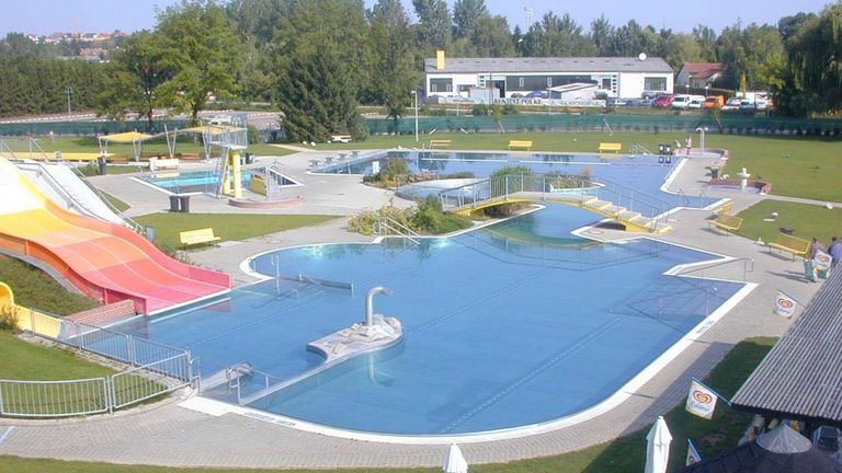 An aerial view of a large outdoor swimming pool with multiple lanes, a diving board, and a yellow bridge connecting the pools. There are several benches and a building in the background.