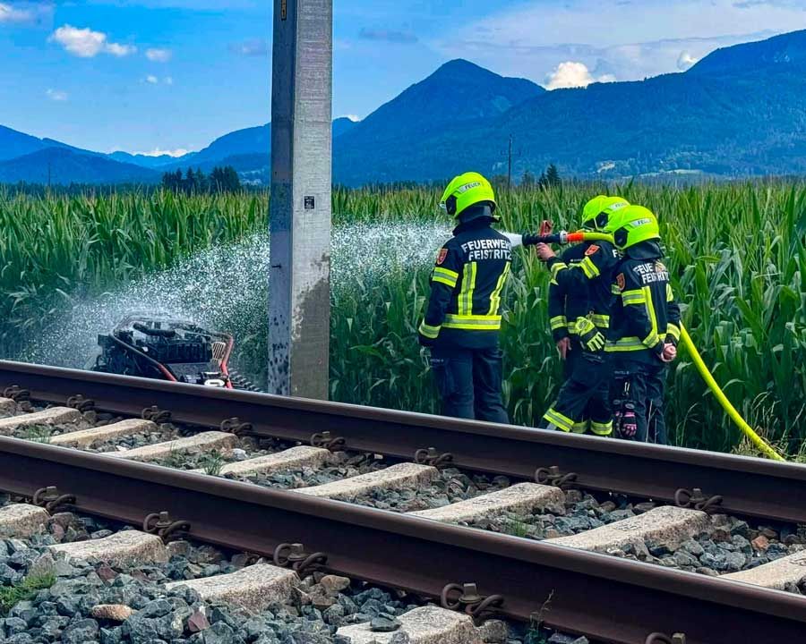 Two firefighters in green uniforms are spraying water from a hose onto a train track. The track is surrounded by tall corn and a pole. In the background, there are mountains.