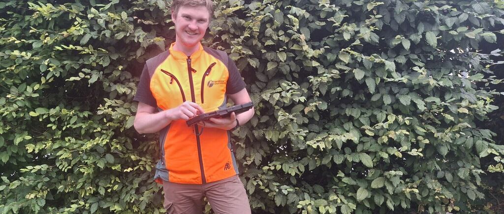 A man in an orange safety vest is standing in front of a bush, holding a device in his hands.