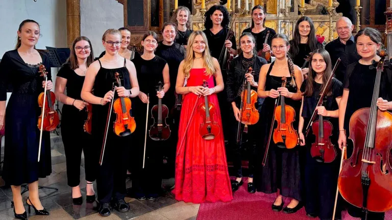 A group of women musicians, dressed in black, are posing for a photo while holding violins. One woman in a red dress stands out in the center.