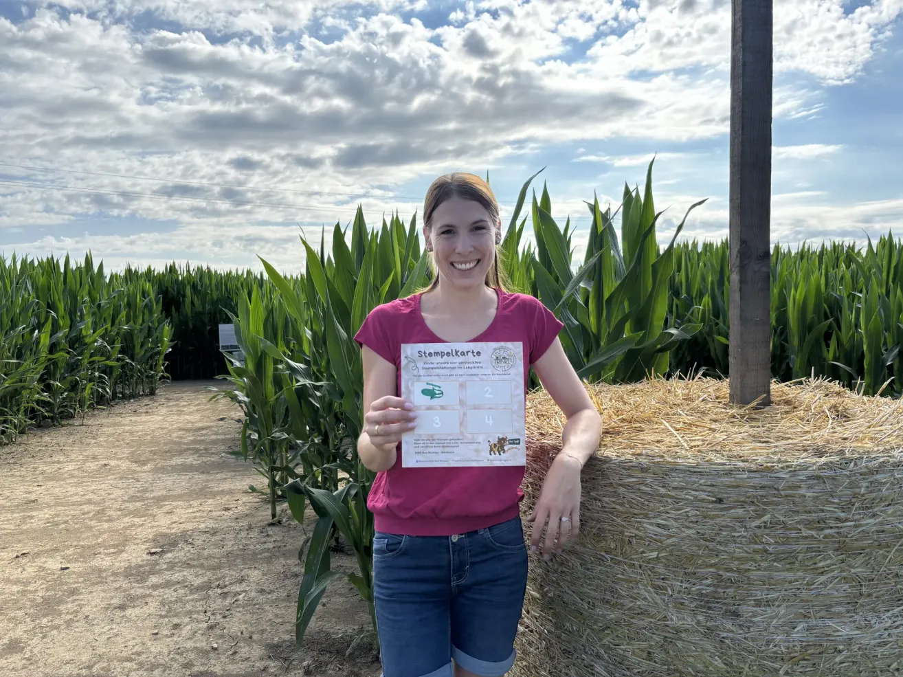 A woman in a pink shirt and blue shorts is standing in a cornfield, holding a stamp card and smiling.