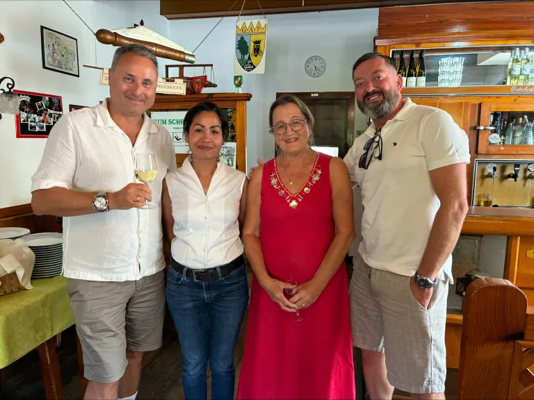 Four adults stand together in a room, smiling for a photo. Two men hold wine glasses, and two women wear glasses and a necklace. A clock and bottles are in the background.