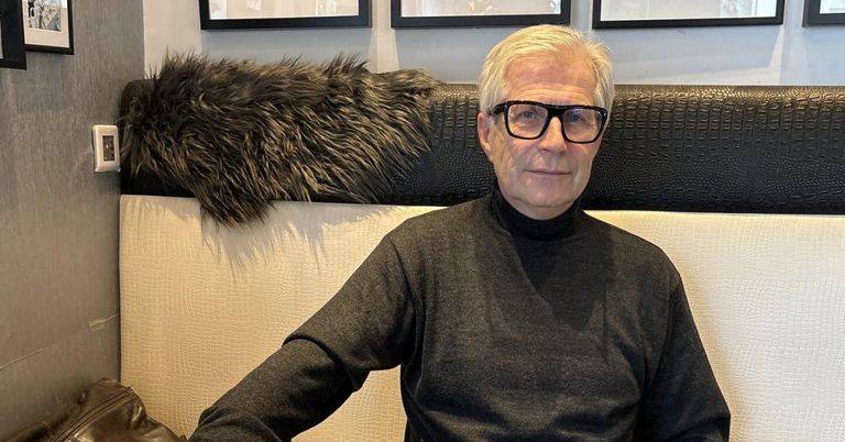 An older man with white hair and glasses sits on a couch with a fur throw over the backrest. He wears a black turtleneck and smiles at the camera.