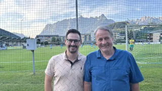 Two men stand in front of a soccer field with mountains in the background. One man wears glasses and a patterned shirt, while the other wears a blue polo.